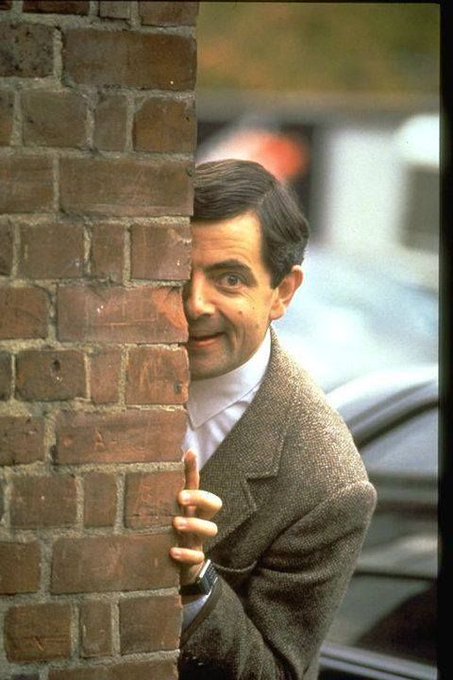 The image features a man peeking out from behind a brick wall, with his hand resting against the wall. The man appears to be smiling or making a face as he looks over the top of the wall. The background shows cars parked on the street, suggesting that this scene takes place in an urban setting.