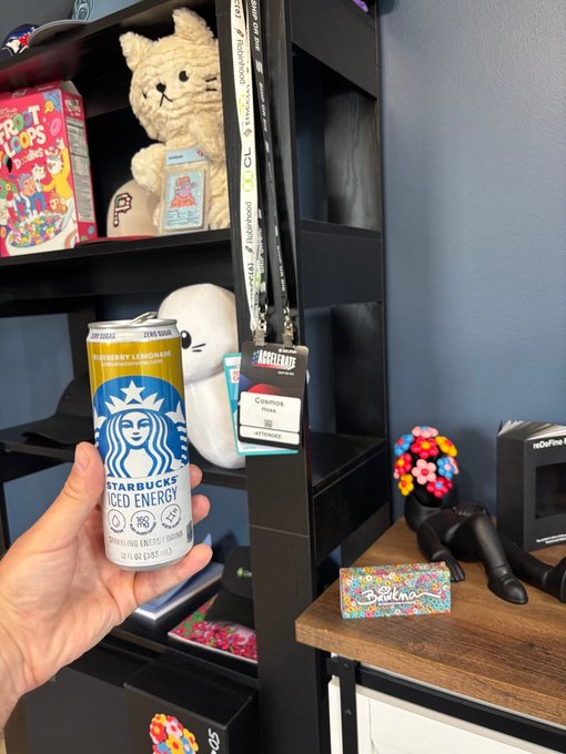 A hand holds a Starbucks Iced Energy Blueberry Lemonade can, with an ID badge reading "Cosmos Hoss" hanging in front of a shelf filled with personal items including a teddy bear, snacks, and gaming accessories—matching the post's Sunday morning greeting to the badge's name.