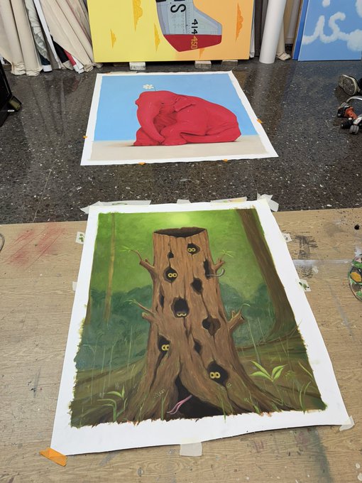 The image features two paintings of trees, one larger than the other. They are displayed on a wooden table with tape covering them up. The larger painting has a tree trunk that appears to be hollow or cut out, while the smaller painting depicts a tree with holes in its bark. Both paintings have a green color scheme and depict forest scenes. The text "GM" is visible in the image as well.