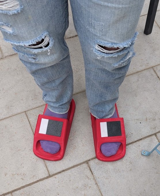 A person wearing ripped blue jeans, purple socks, and red slides with a black and white logo stands on a tiled floor. The casual, relaxed attire serves as a humorous response to the question about their profession, implying a laid-back lifestyle or a job that doesn't require formal clothing.