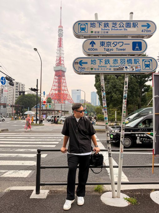 A person in a black shirt and sunglasses stands at a crosswalk near Tokyo Tower, next to bilingual directional signs pointing to Subway Shibakoen Sta. (470m), Tokyo Tower (400m), and Subway Akabanebashi Sta. (70m).