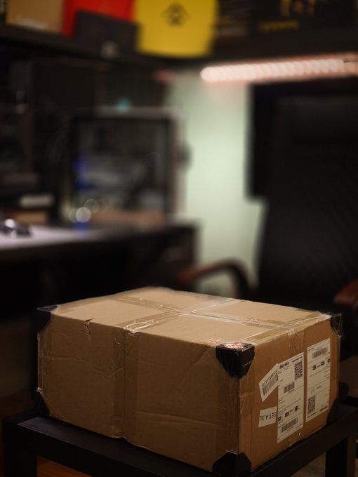 A brown cardboard shipping box sealed with tape and shipping labels sits on a black stool in a dimly lit room, teasing an upcoming reveal to the Binance community.

The labels include numbers like "3051 0055 2902 1" and a tracking number "5841 9001 3128 9358 6895 55," indicating the package is freshly delivered and unopened.