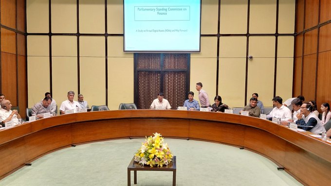 Members of India's Parliamentary Standing Committee on Finance are seated around a large circular table during a meeting, with a presentation screen in the background displaying the committee's name and its focus on crypto regulation, as they take oral evidence from the Finance Ministry.