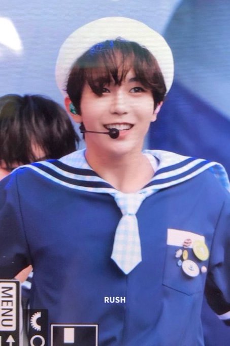 A K-pop performer wearing a sailor-style blue outfit and white sailor hat smiles into a microphone headset during a stage performance, with the word 'RUSH' visible at the bottom and 'MENU' on the side.