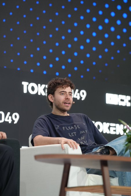 A young man with curly brown hair and a beard, wearing a dark t-shirt that reads "PLEASE LET AN AI PAY," sits on stage at what appears to be a TOKEN2049 conference event.