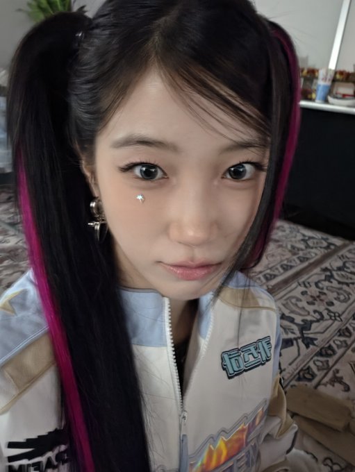 A close-up selfie of a young woman with black pigtails featuring pink streaks, wearing dramatic makeup with a small rhinestone under her eye and a patterned jacket.
