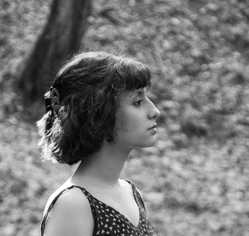 A black and white profile portrait of a young woman with curly, short hair, wearing a polka-dot top, set against a softly blurred natural background.