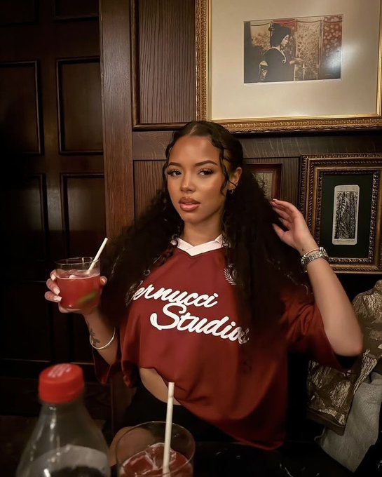 A young woman with long dark hair wearing a maroon 'Marucci Studio' top, holding a red cocktail in a dimly lit bar with framed artwork on dark wood paneled walls.