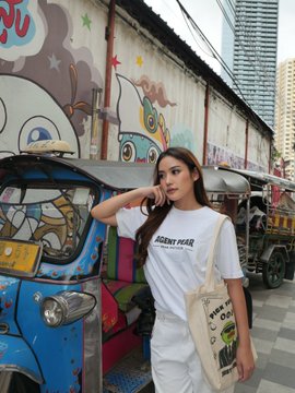A woman wearing a white 'Resident Hero' t-shirt and white pants stands in front of a graffiti-covered wall and a blue tuk-tuk in an urban setting.