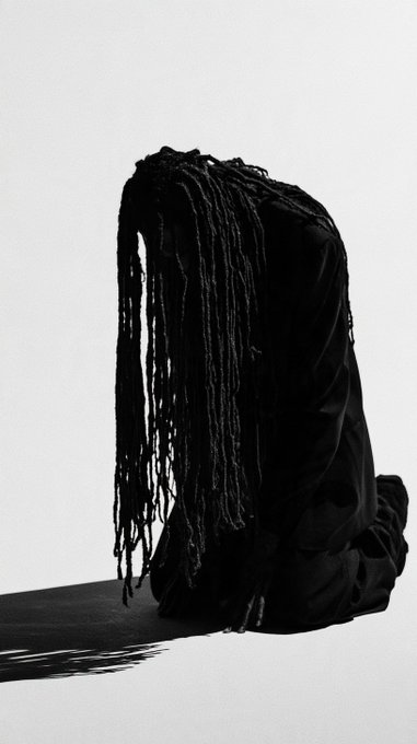 A striking artistic black-and-white photograph of a figure with long dark dreadlocks wearing flowing black fabric, posed against a plain light background with a dramatic shadow cast on the floor.