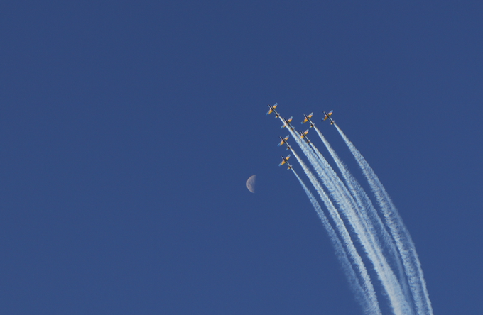A formation of jet aircraft flies in tight formation through a clear blue sky, leaving long white contrails behind them, with a small crescent moon visible in the background.