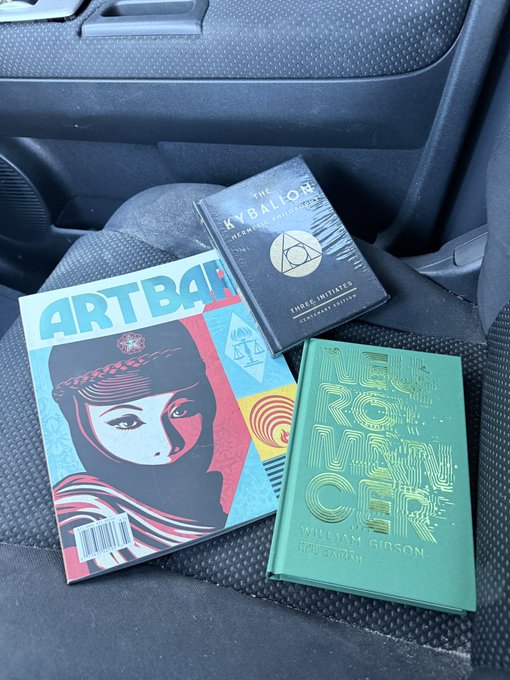 Three books rest on a car seat: a Shepard Fairey "ARTBAHN" paperback, "The Kybalion: Hermetic Philosophy Anniversary Edition," and William Gibson's "Neuromancer" (signed edition), representing the creator's haul from a research trip to Barnes & Noble after missing sought-after trading cards.