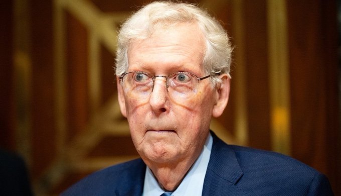A close-up portrait of an elderly man with white hair and glasses, wearing a dark blue suit jacket over a light blue shirt.