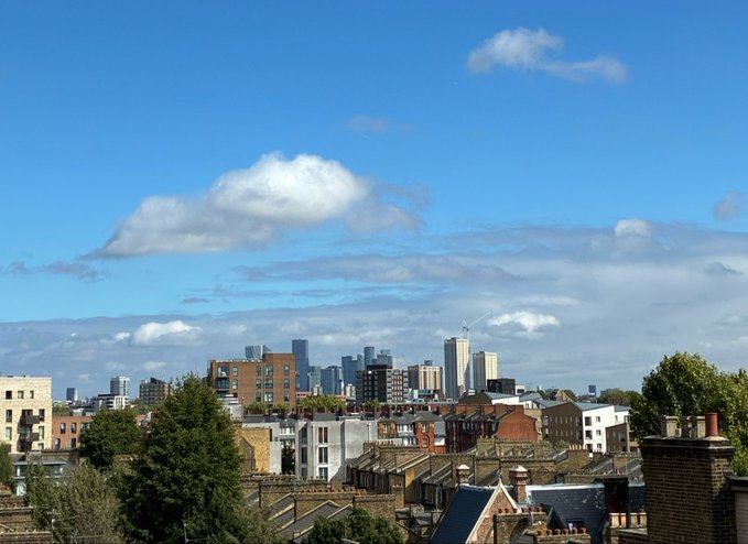 A cityscape view showing a skyline with skyscrapers in the distance behind residential rooftops and trees under a bright blue sky with scattered clouds.