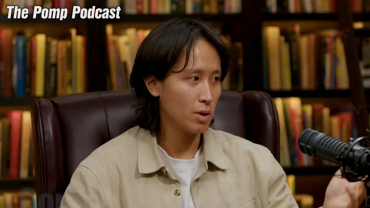 The image features a man sitting in a chair, wearing a tan jacket, with his head tilted back as if he's deep in thought or contemplation. The background of the image shows numerous bookshelves filled with various books, suggesting that this scene takes place in a library or a reading area. The caption for the image reads: "The Pimp podcast is being interviewed by @Rewkang on the topic 'Interesting watch'. Focus on what is in the image and how it relates to the post."