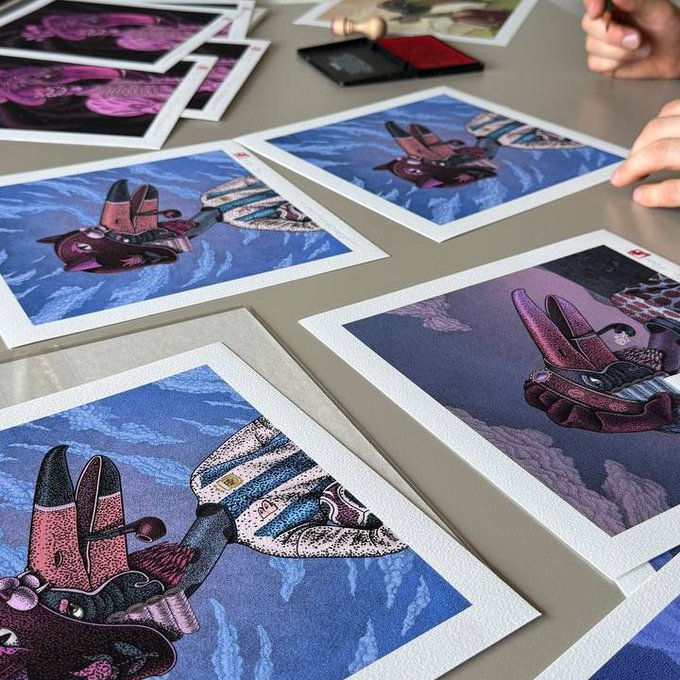 Photograph of multiple colorful, whimsical art prints featuring creature-like characters with airplane wings laid out on a gray table, with an ink stamp and hands visible in the background.