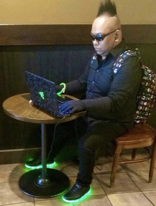 A man with a mohawk and sunglasses sits at a small table using a glowing green Razer laptop, wearing black gloves and a studded backpack, with LED lights glowing under his shoes.
