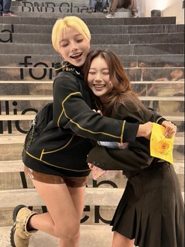 Two young women share a happy hug on a staircase; one has blonde hair and wears a black jacket, while the other has brown hair and holds a yellow envelope.