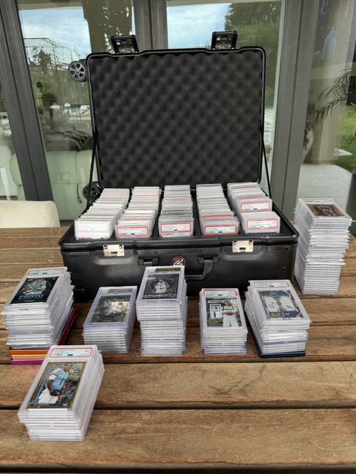 An open black hard case filled with stacks of graded sports cards in plastic sleeves sits on a wooden table, with many more graded card stacks arranged in front of it.