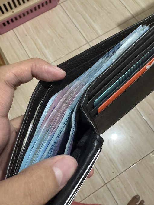 A hand holds an overstuffed black wallet crammed with various banknotes and colorful cards, visually echoing the wish that wallets stay full and prosperity abounds. Partial text on one of the bills reads "LIMA MONEDA."