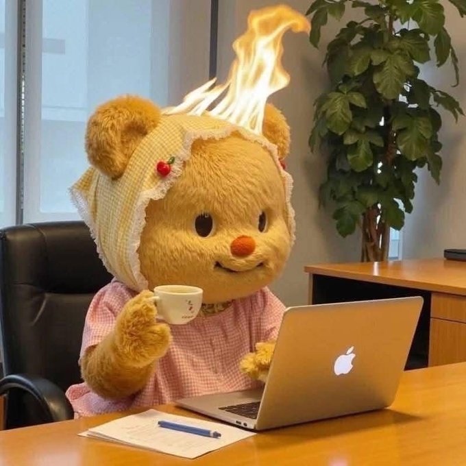 A bear mascot dressed in a pink checkered dress and bonnet with a cherry accent sits at a desk, sipping from a small teacup while flames blaze on top of its head, working on a silver Apple laptop with a notebook and pen nearby.