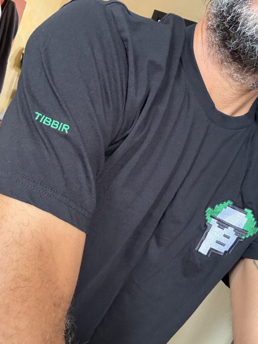 A close-up photo of a man wearing a black t-shirt featuring a pixel-art graphic of a block with green foliage and the word 'TIBBIR' printed in green on the sleeve.
