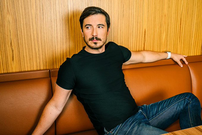 Robinhood CEO Vlad Tenev is shown in a relaxed pose, wearing a black t-shirt and jeans while sitting against a wood-paneled wall on a tan leather booth. The image accompanies his call for U.S. policymakers to embrace tokenized stocks, highlighting his role as a fintech leader advocating for innovative trading solutions.