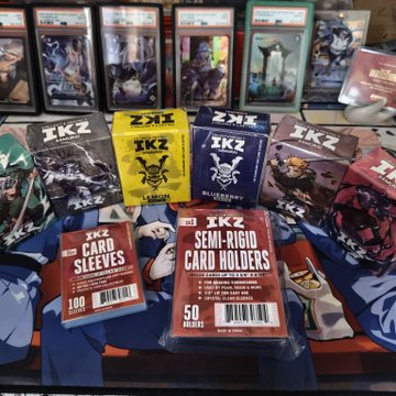 A display of IKZ Samurai trading card game merchandise, including booster boxes and various card sleeves (card sleeves shown in packs of 100, with semi-rigid holders in packs of 50). The winner proudly showcases their prize haul from @azukirunclub, highlighting that while the transparent sleeves aren't tournament legal, the Rose and Blueberry sleeves are.
