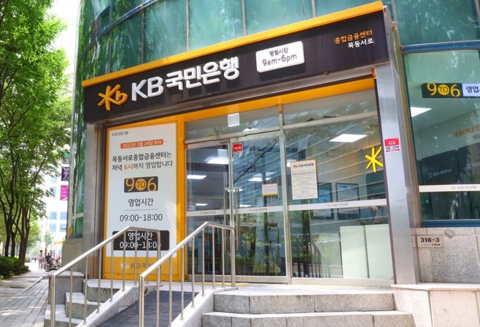 The storefront of Kookmin Bank (KB국민은행) in Korea is shown, with signage indicating business hours of 9am-6pm and phone number 906. The post draws a parallel between Kookmin Bank's adoption of JP Morgan's blockchain technology and Bitcoin's cumulative positives, framing institutional blockchain integration as another positive signal for Bitcoin's continued growth.