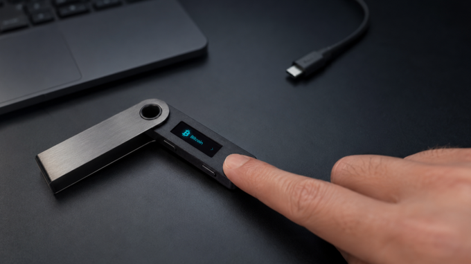 A close-up product photo of a person using their finger to press a Ledger hardware wallet, which displays 'Bitcoin' on its small screen, connected via USB to a laptop on a dark desk.