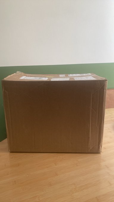A sealed brown cardboard shipping box with shipping labels sits on a bamboo wooden floor against a green and white wall.