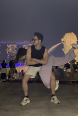 A young man wearing glasses, a black tank top, white shorts, and white sandals crouches on a rooftop at dusk, pointing his finger toward the camera with a harbor and city skyline visible in the background.