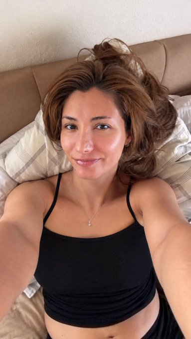 A selfie of a woman with brown hair lying on a bed, wearing a black top and a delicate silver necklace.