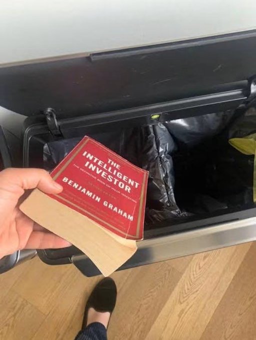 A hand holds a red copy of "The Intelligent Investor" by Benjamin Graham above a trash bin lined with a black bag, with a foot in a black sock visible on the wooden floor below — humorously framing the book being discarded after watching a meme-stock-style fortune flip.