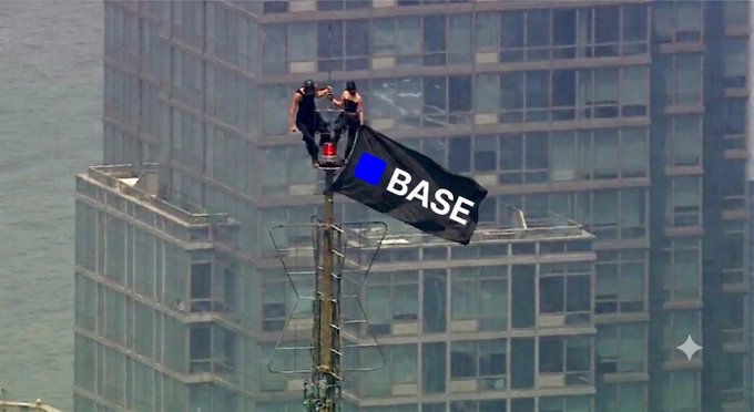 Two workers on a tall antenna or mast attaching a black flag with the 'BASE' logo (blue square) near the top of a skyscraper.