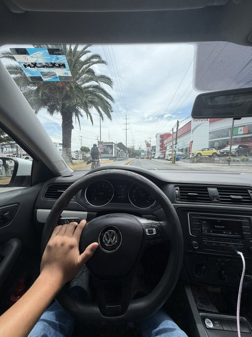 A first-person view from inside a Volkswagen car, with the driver's hand on the steering wheel, looking out at a tropical street with palm trees and a Toyota dealership visible in the distance.