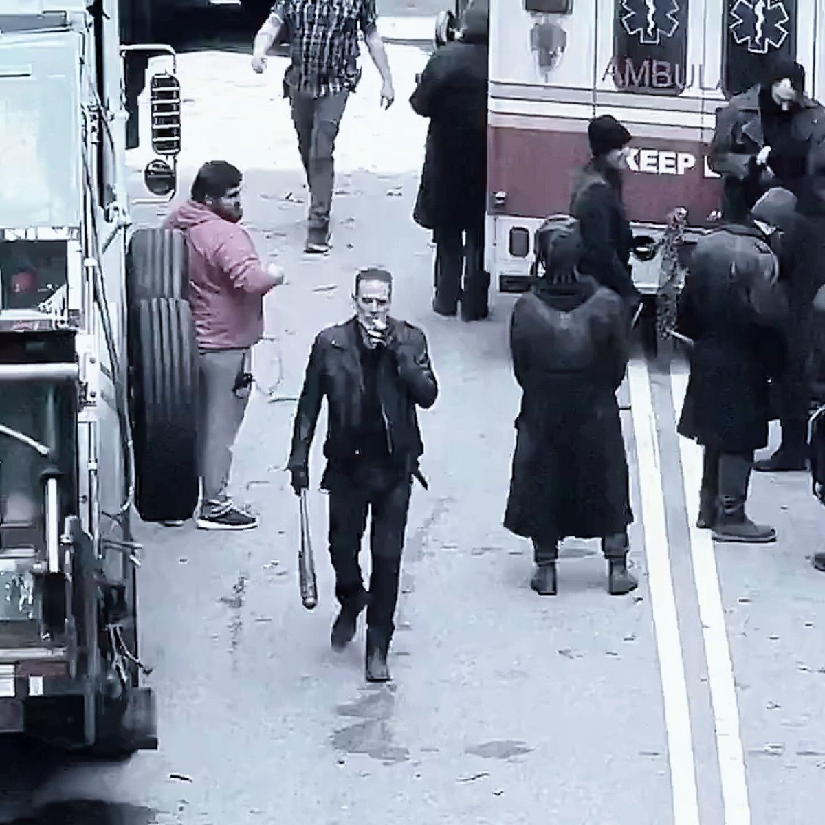 The image shows a man walking down a street, holding a gun. There are several other people around him, some of whom appear to be carrying backpacks or handbags. The scene takes place near an ambulance, suggesting that there might have been an emergency nearby. The photo is taken from an elevated perspective, providing a clear view of the entire area and the individuals present in it.