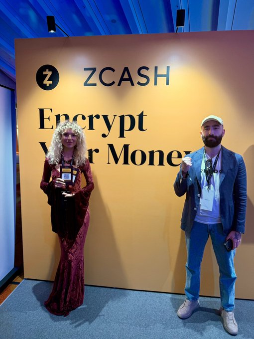 Two people standing in front of a large yellow ZCASH advertisement banner that reads 'Encrypt Your Money,' both wearing event lanyards and holding items in their hands.