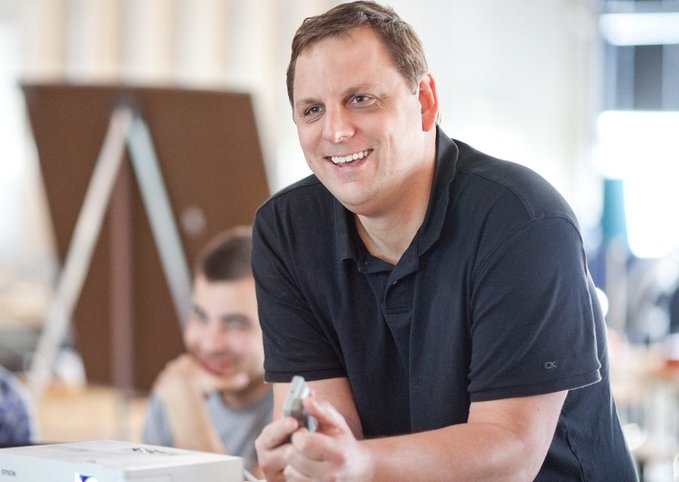The image shows a smiling man in a dark polo shirt holding what appears to be a small object, with another person blurred in the background. There is no visible text, numbers, tickers, or names in the image itself, so no such details can be transcribed. The photo appears to be a generic stock-style image of a professional man, which does not specifically depict Michael Arrington, XRP, hedge funds, or any of the post's claims about institutional crypto infrastructure.