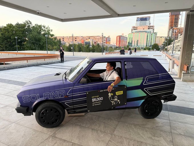 A person holds a sign reading "claiming $5,000" while seated in a car wrapped in Solana branding and graphics in an urban plaza. The "wagmi" caption reflects the celebratory crypto giveaway moment, suggesting we're all going to make it together.