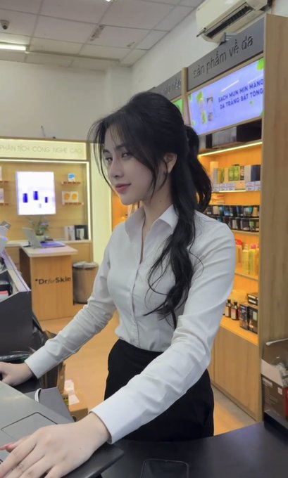 A young woman with long dark hair, wearing a white shirt and black skirt, stands at a counter in what appears to be a beauty or skincare store, with Vietnamese signage visible on the shelves behind her.