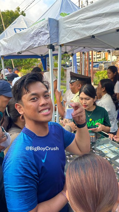 The image shows a smiling young man in a blue Nike shirt at an outdoor market stall under white tents, with people gathered around what appears to be a gold-buying event. The watermark "@BigBoyCrush" is visible on the shirt, connecting to the post's playful caption about "snacking on food" and "snacking on gold," suggesting the setting is likely a gold-buying or appraisal event where people can exchange items for cash.