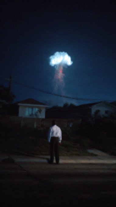 A man in a white shirt and dark pants standing on a dark street at night, looking up at what appears to be a nuclear mushroom cloud explosion in the sky above a residential neighborhood.