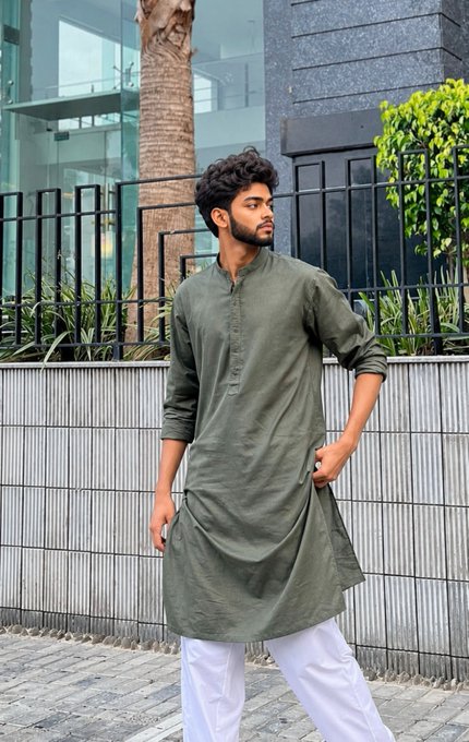 A young man wearing an olive green traditional kurta and white pants stands outdoors near a metal fence and palm trees.