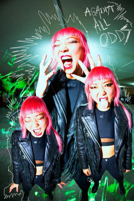Three identical women with pink hair wearing black leather jackets posing energetically, with 'AGAINST ALL ODDS' written in the upper right against a green-lit background.