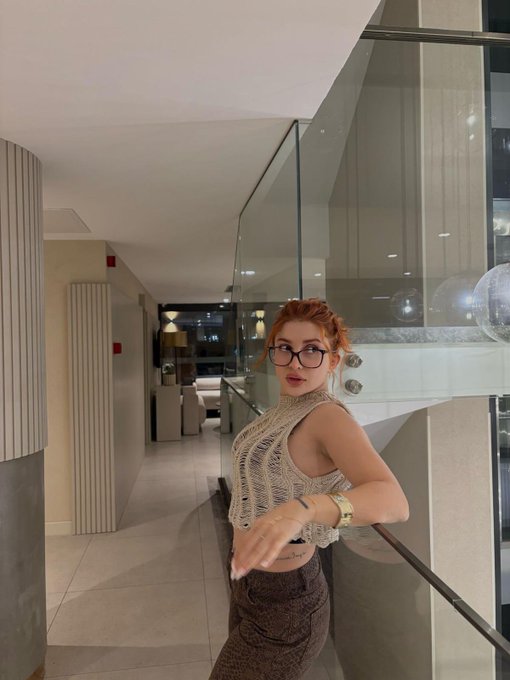 A woman with red hair, glasses, and a knit halter top stands in a modern interior featuring glass walls and curved architectural details. The image's calm, confident pose and upscale setting visually echo the post's message about patience and restraint in crypto investing—suggesting that composure and stillness, rather than constant action, are key to long-term success.