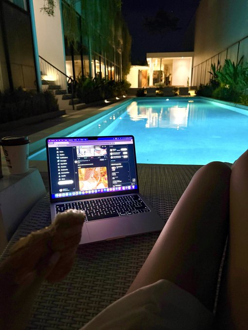 A nighttime scene by a pool with a laptop and coffee cup on a lounge chair, with a cat's paw visible in the foreground, showing an image editing software on the screen.