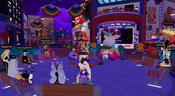 A vibrant virtual plaza in Decentraland, decorated with rainbow pride flags, features various avatars (including usernames like "Kenzo," "ToxicWaifu," and "darkcity") gathered around benches and a large screen displaying "Live Events." The neon-lit nighttime scene captures the social, immersive atmosphere of a digital hangout space.