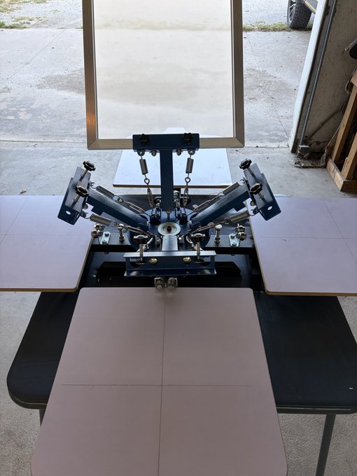 A four-color manual screen printing press sits on a worktable with three blank platens ready for printing.