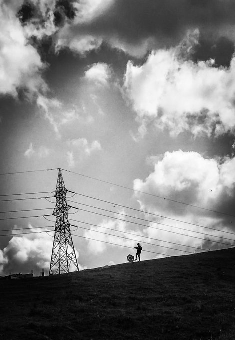A black and white photograph showing a silhouetted figure walking on a hill carrying a ball near a power transmission tower under dramatic cloudy skies.
